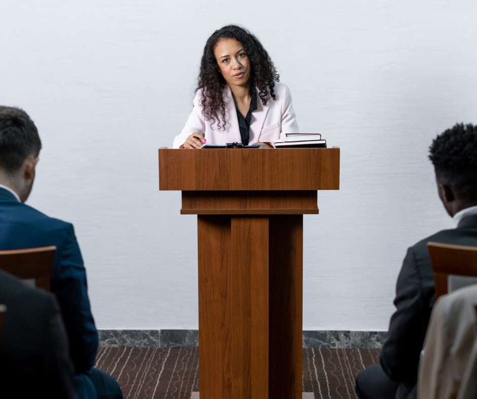 A a photo of a woman standing at a wooden pulpit, speaking.